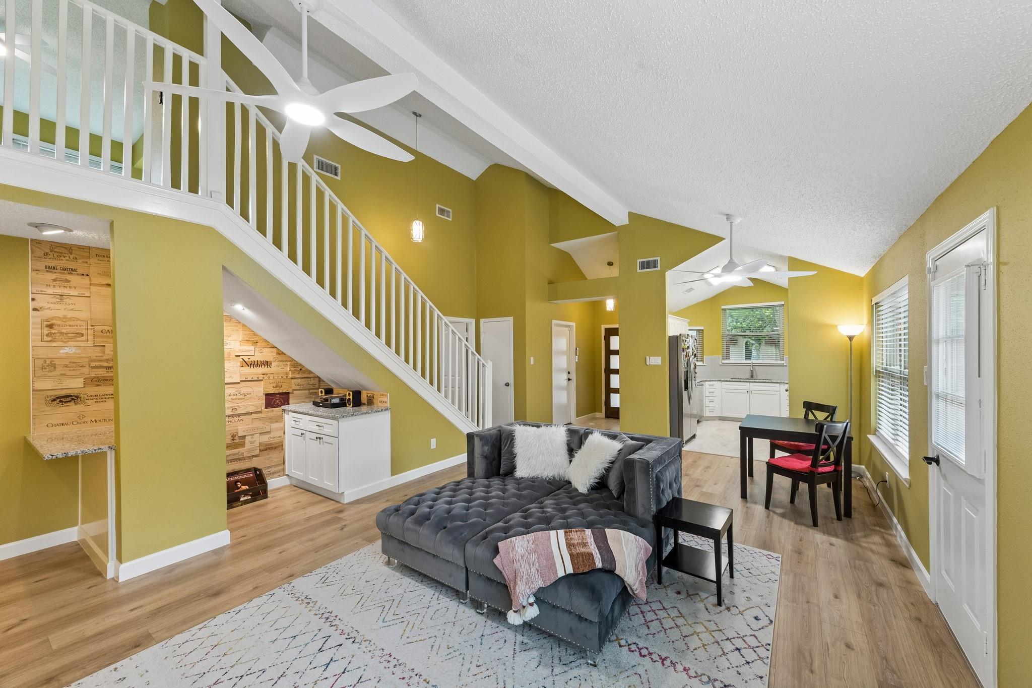 Open-plan living room with a gray sectional beneath a white staircase and the kitchen beyond.