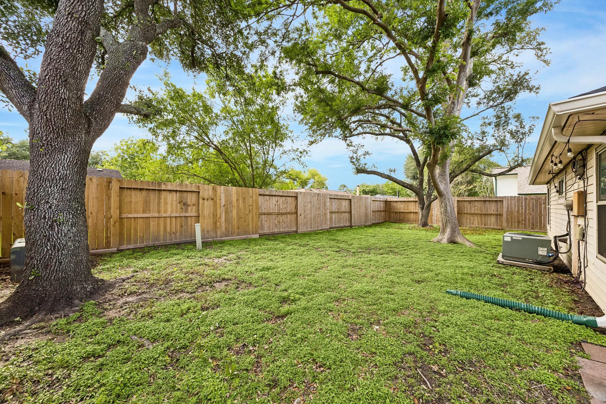 Fenced backyard with a green lawn, mature shade trees, and the house exterior.