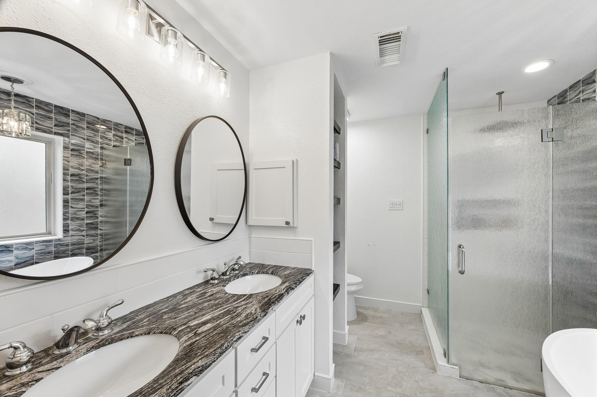 Bathroom with a double-sink vanity, veined countertop, round mirrors, glass shower, and bathtub.