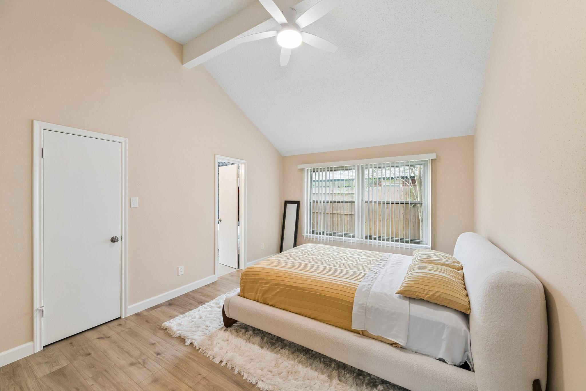 Staged bedroom with vaulted ceiling, mustard-gold bedding, and a wide window with vertical blinds.
