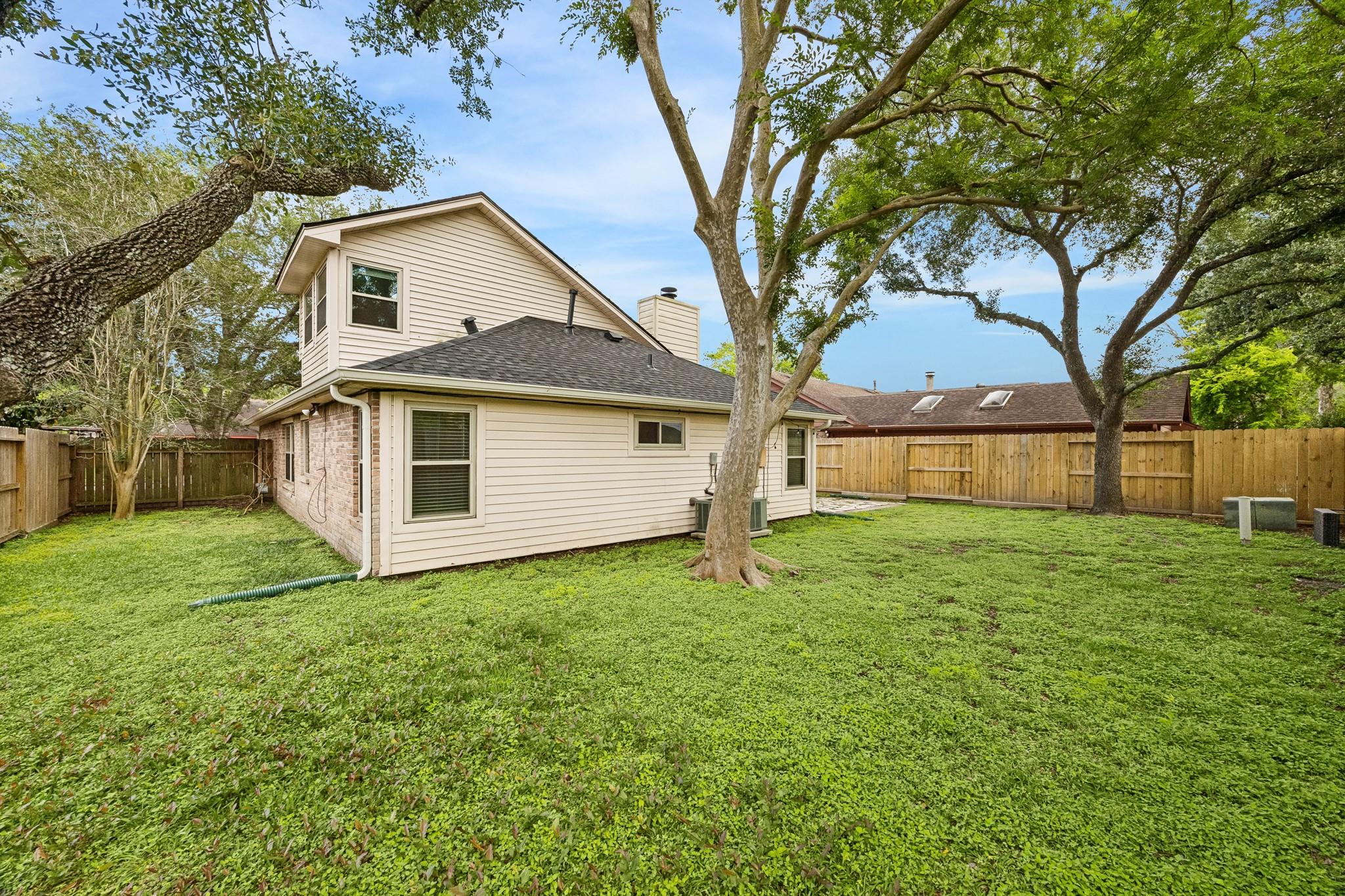 Rear of a beige two-story home behind a large grassy yard shaded by mature trees.