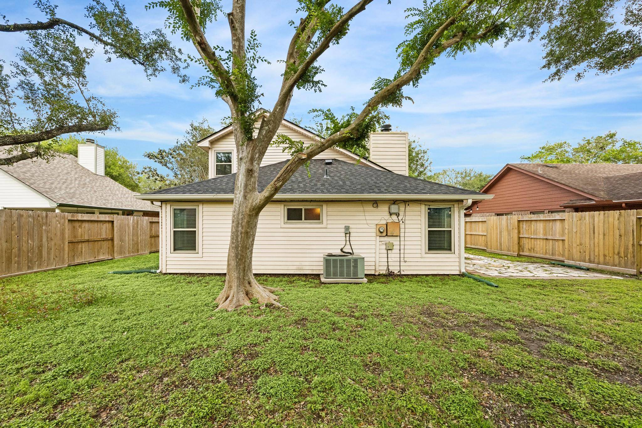 Rear of a light-colored two-story house behind a large tree in a fenced grassy yard.