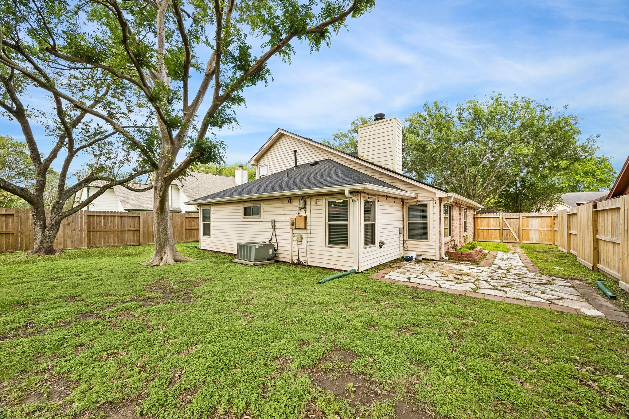 Rear of a light-colored house with a stone patio, fenced yard, and mature trees.