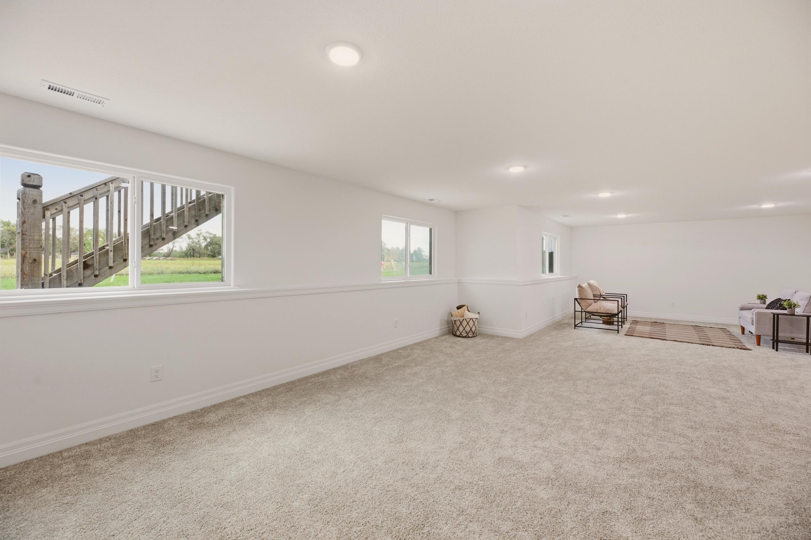Spacious carpeted basement living area with wide windows and a small seating area.