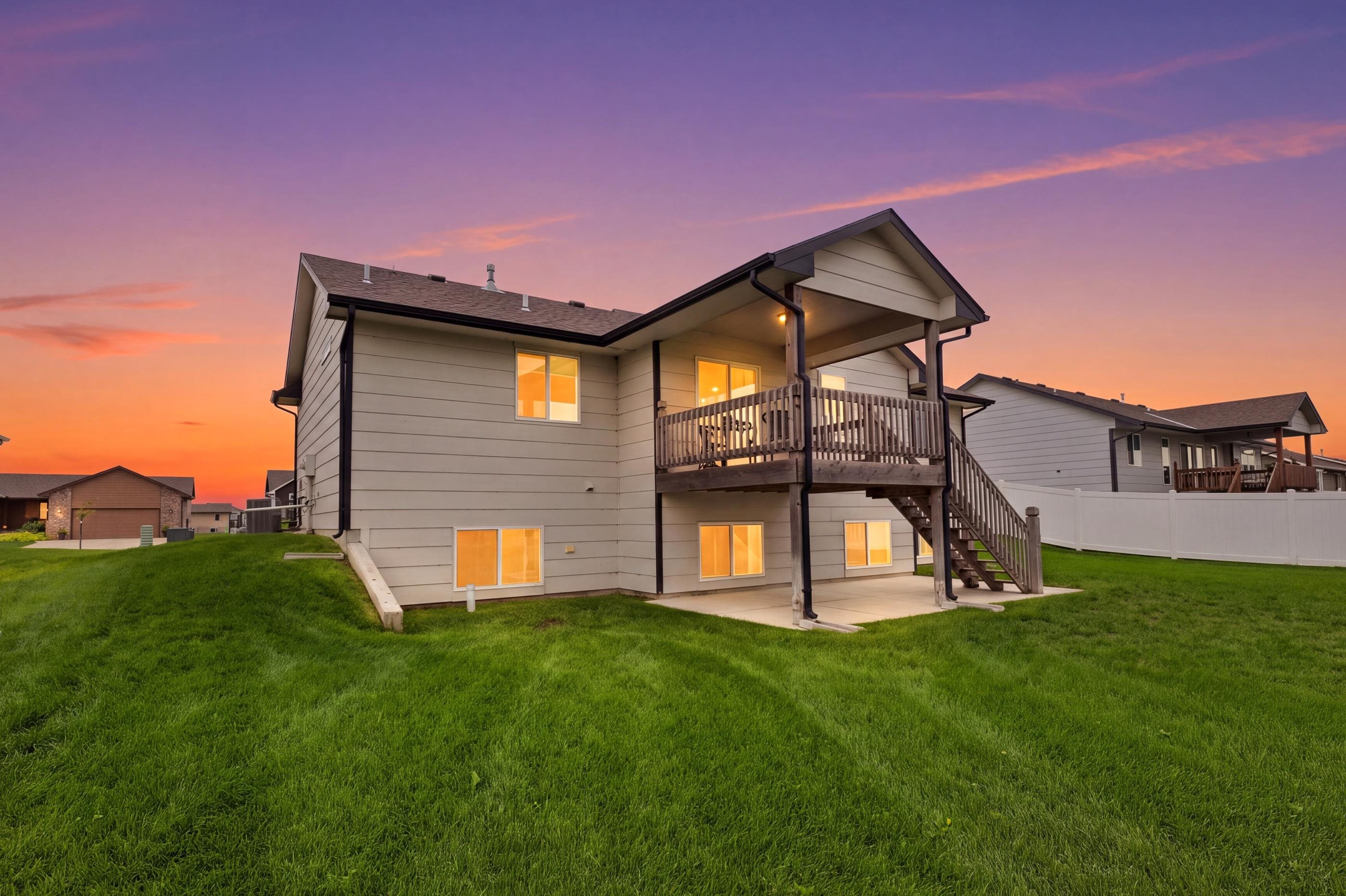 Home’s rear exterior at sunset, with a raised wooden deck, stairs, and illuminated walkout lower level.