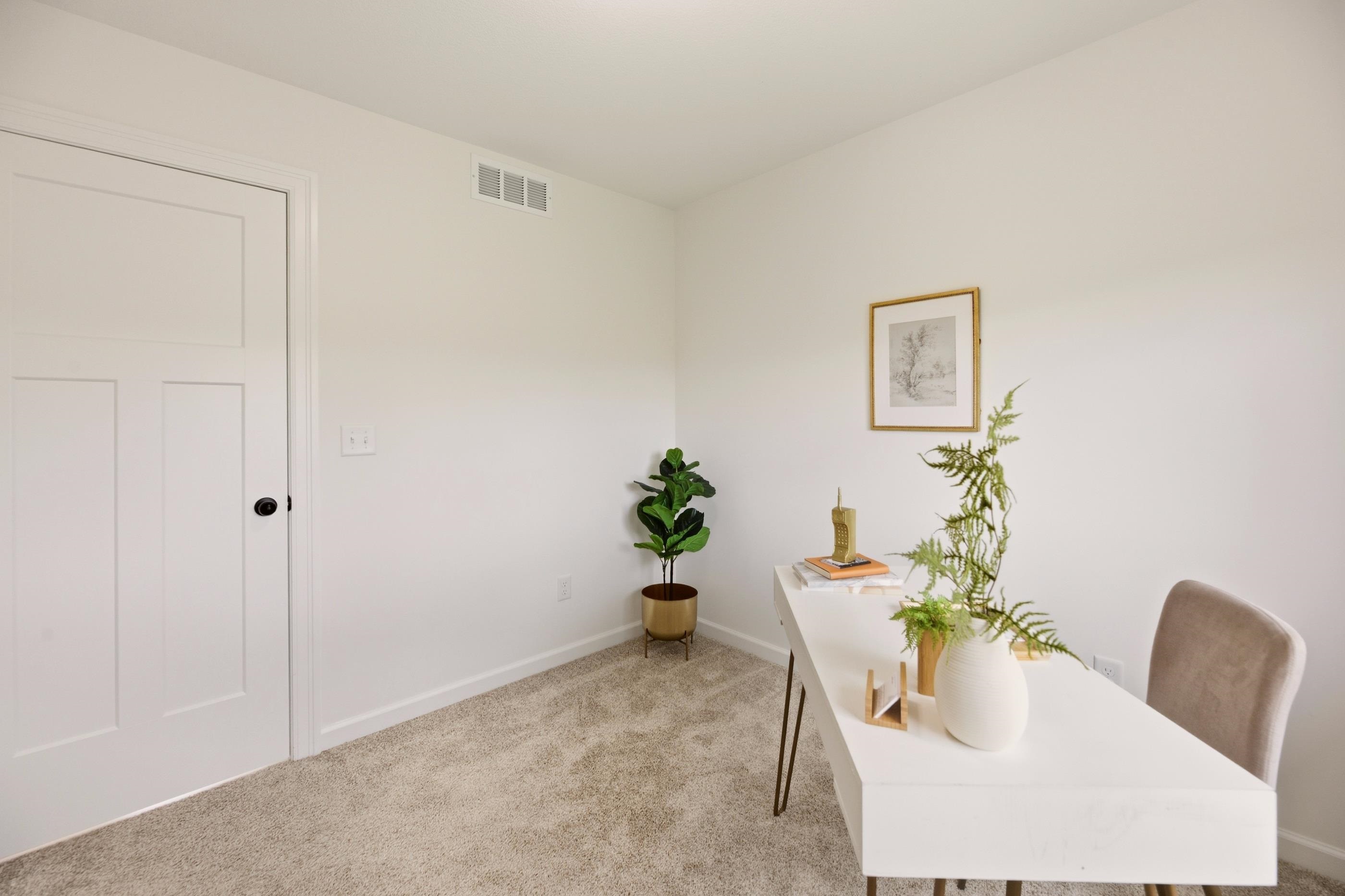 Staged home office with a white desk, beige carpet, framed art, and potted plants.
