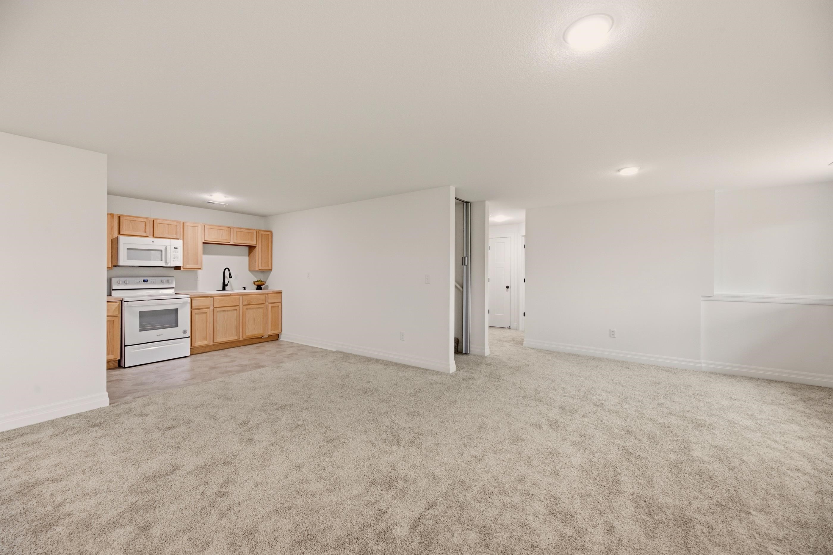 Unfurnished carpeted living area with a compact light-wood kitchenette and white appliances.
