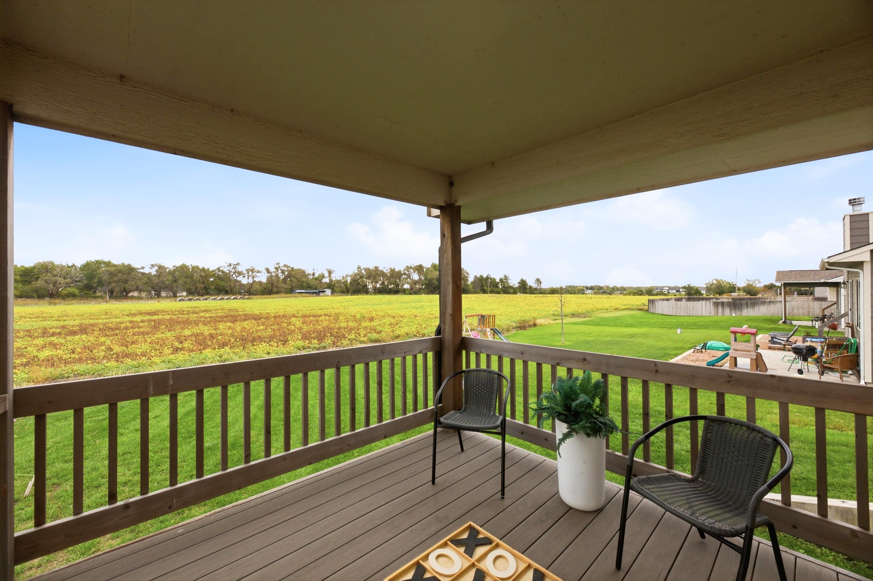 Covered deck overlooks a green lawn, children’s play area, and yellow field.