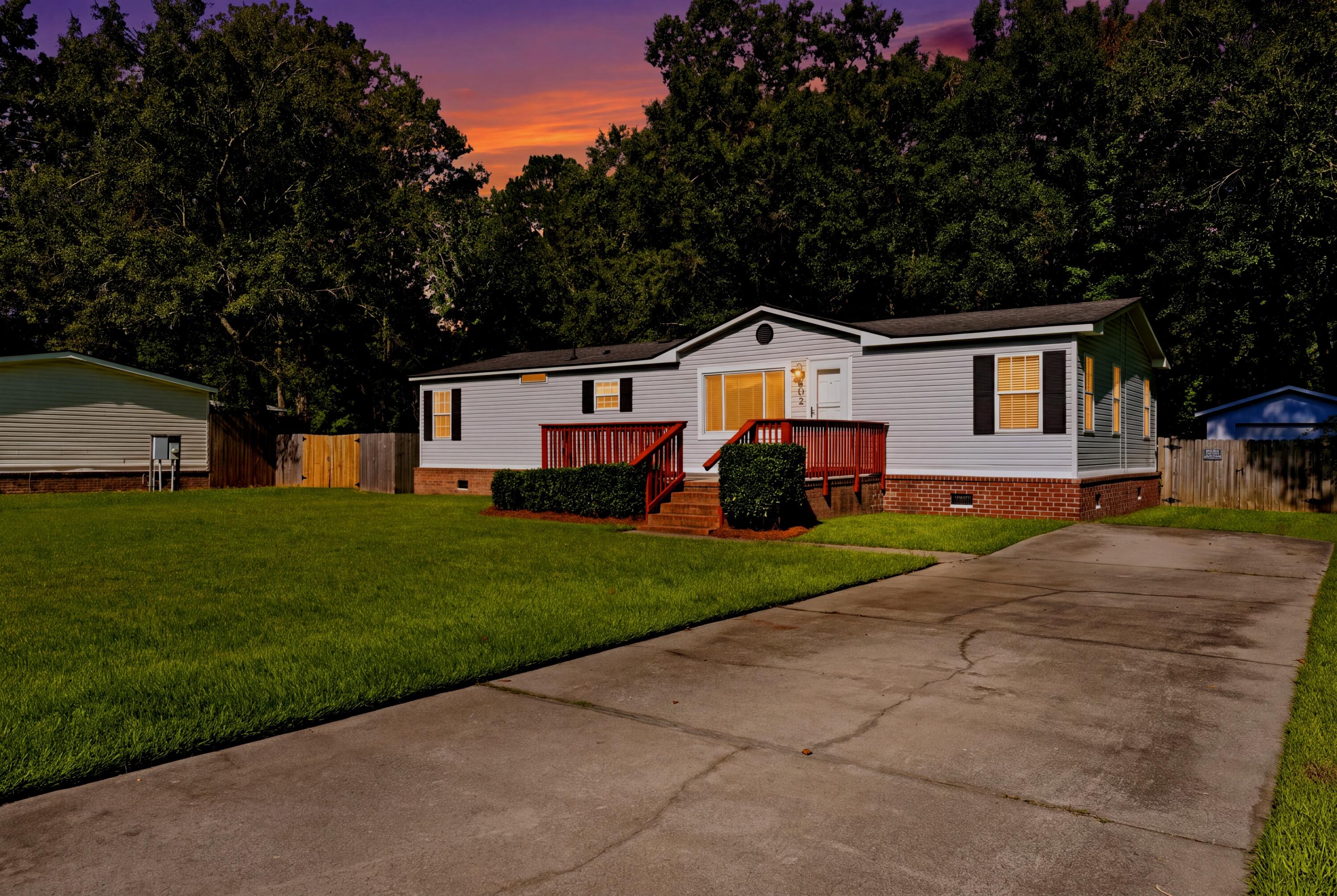 White-sided home with a red-railed porch, brick foundation, broad driveway, and wooded backdrop at dusk.