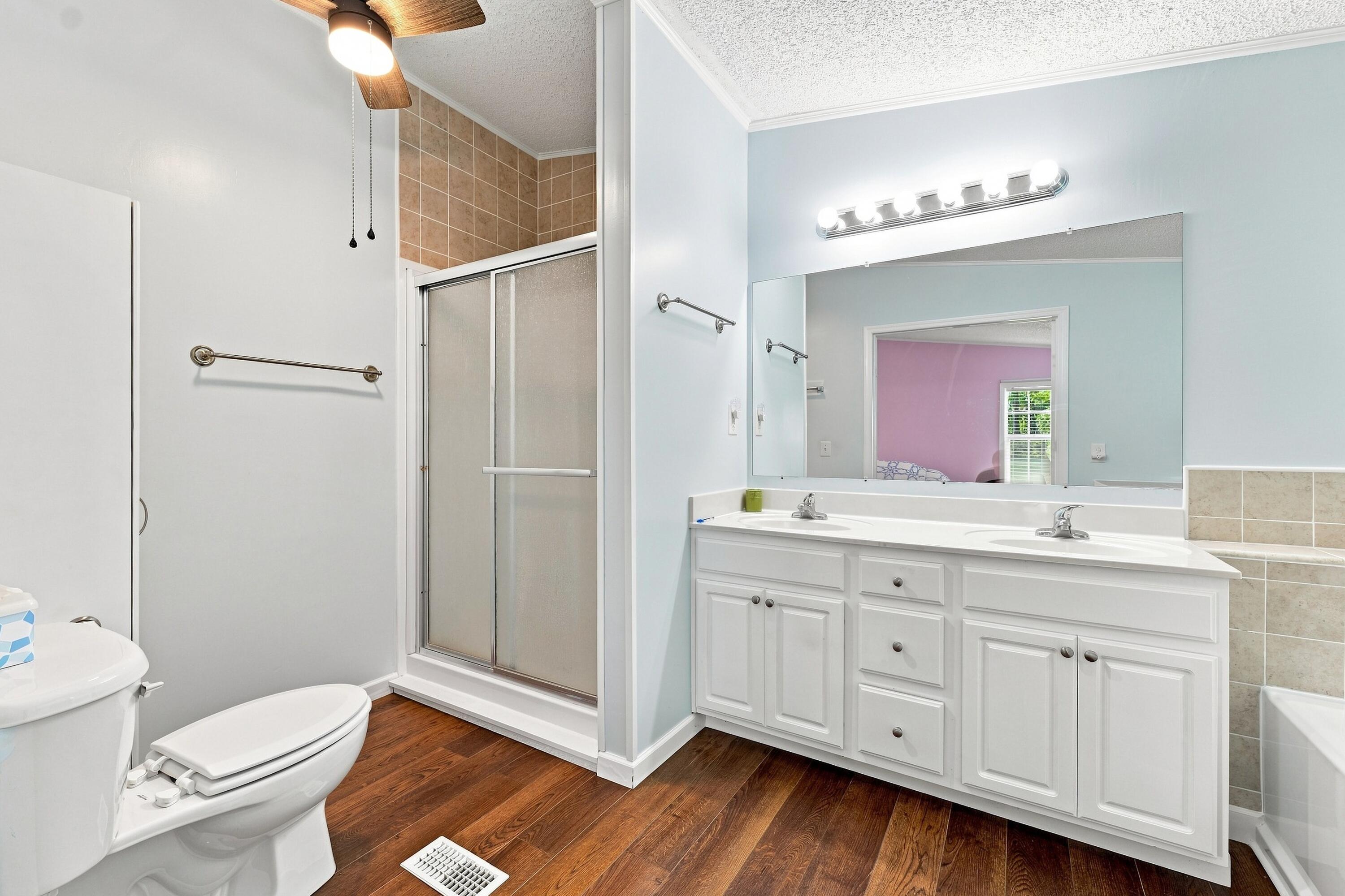 Bathroom with a white double-sink vanity, large mirror, glass shower, and dark wood-look flooring.