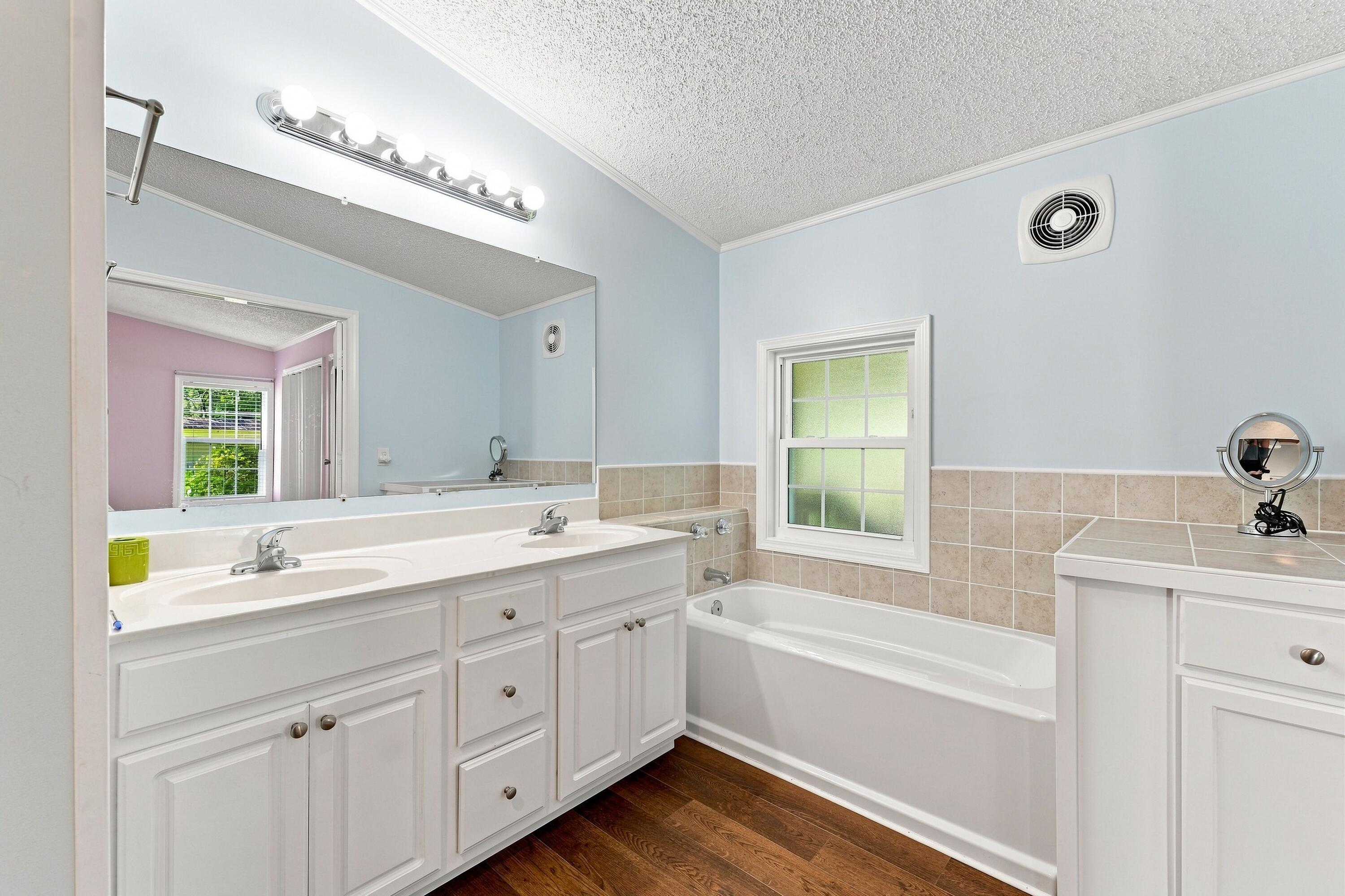 Bathroom with double vanity, bathtub, frosted window, and dark wood flooring.