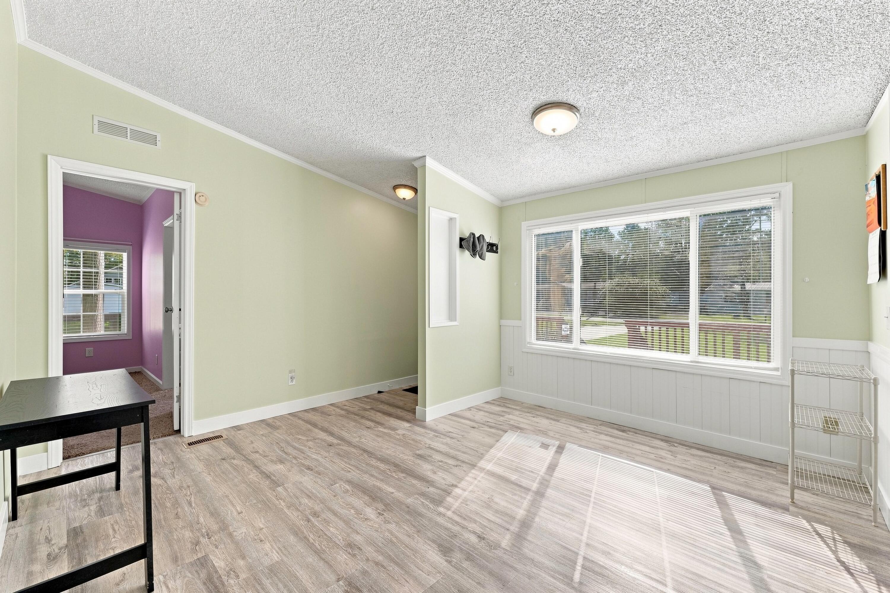 Bright room with gray wood-look floors and a large window overlooking the yard; doorway opens to a purple room.