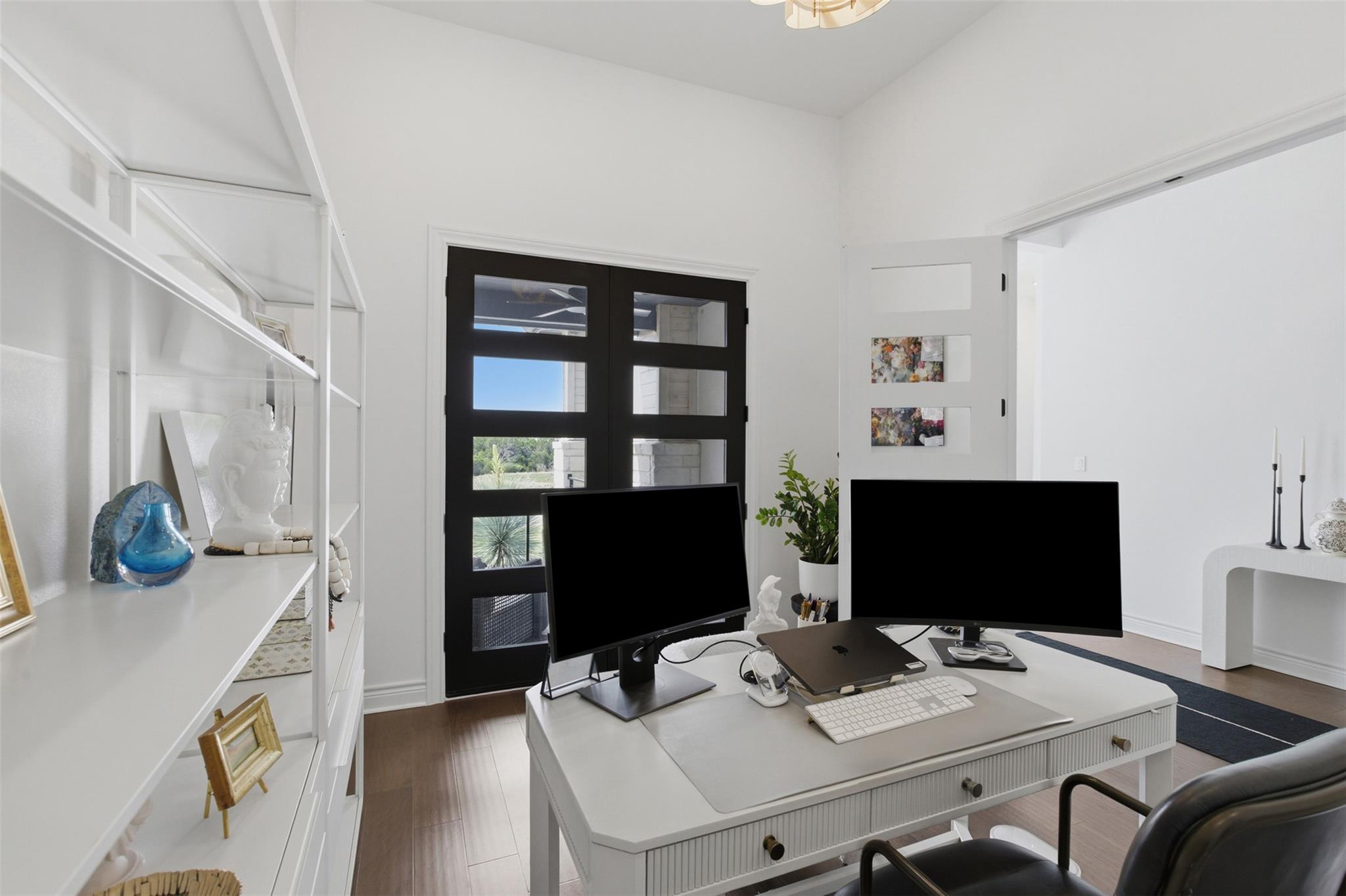 Furnished home office with a white desk, dual monitors, and black-framed glass doors.