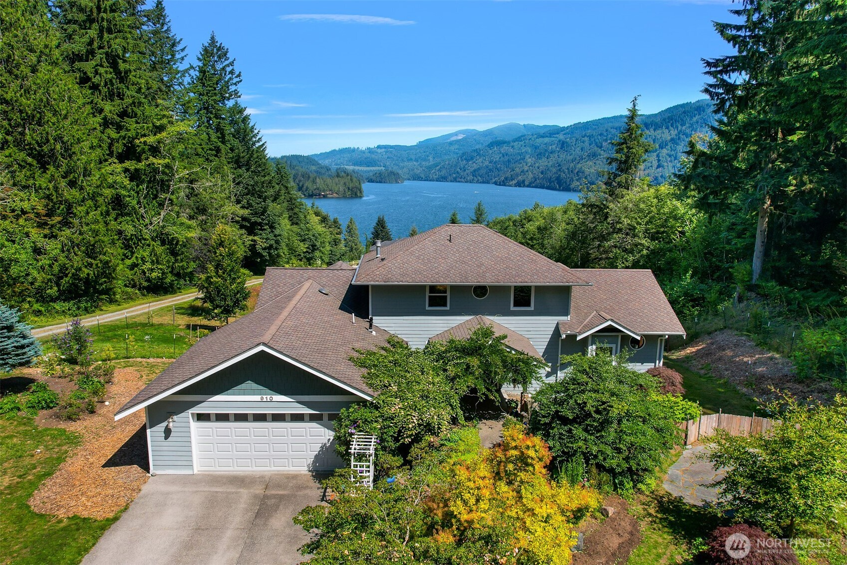 Aerial view of a gray house above a lake, surrounded by evergreens and forested mountains.