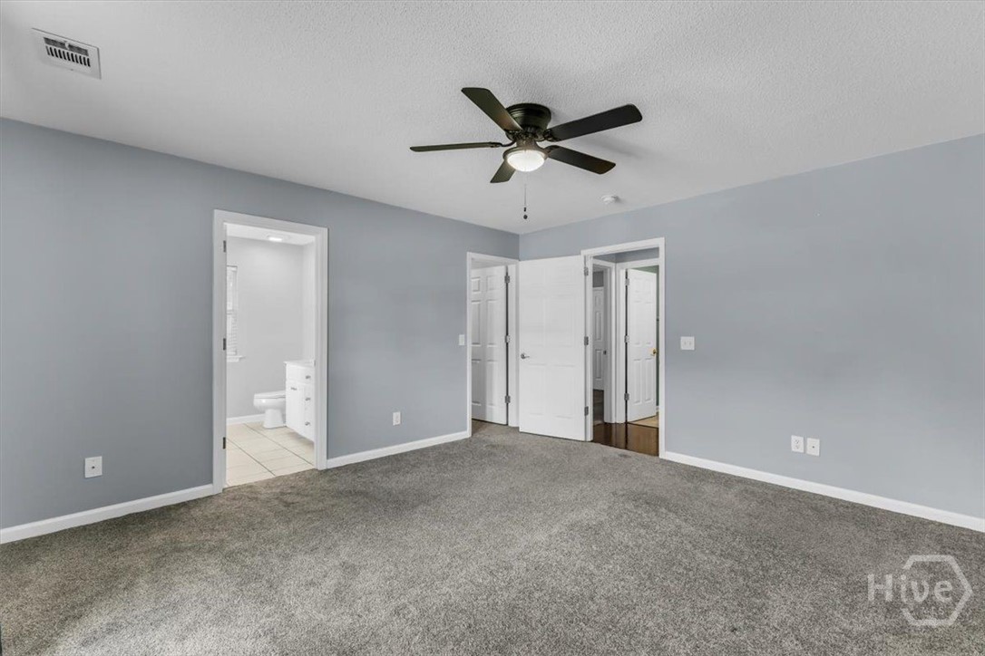 Empty bedroom with gray carpet, ceiling fan, and open doorways to a bathroom and hall.