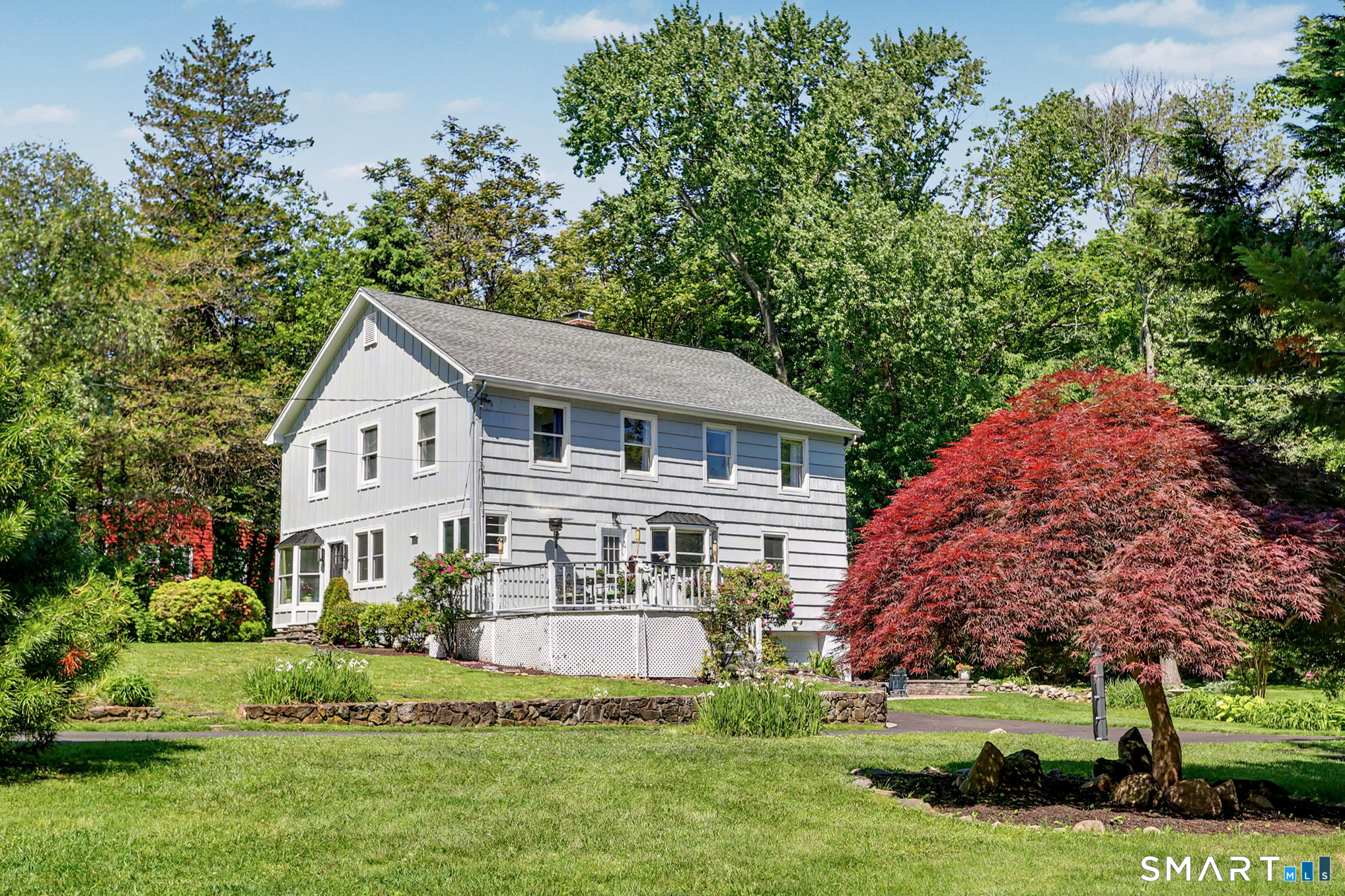 Two-story gray-and-white house with a raised deck, landscaped lawn, and red Japanese maple.