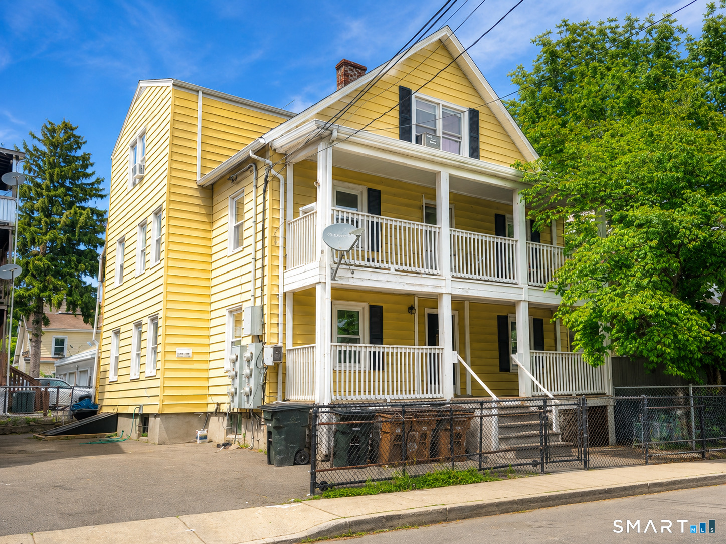 Yellow multi-story house with white stacked porches, chain-link fence, and leafy trees.