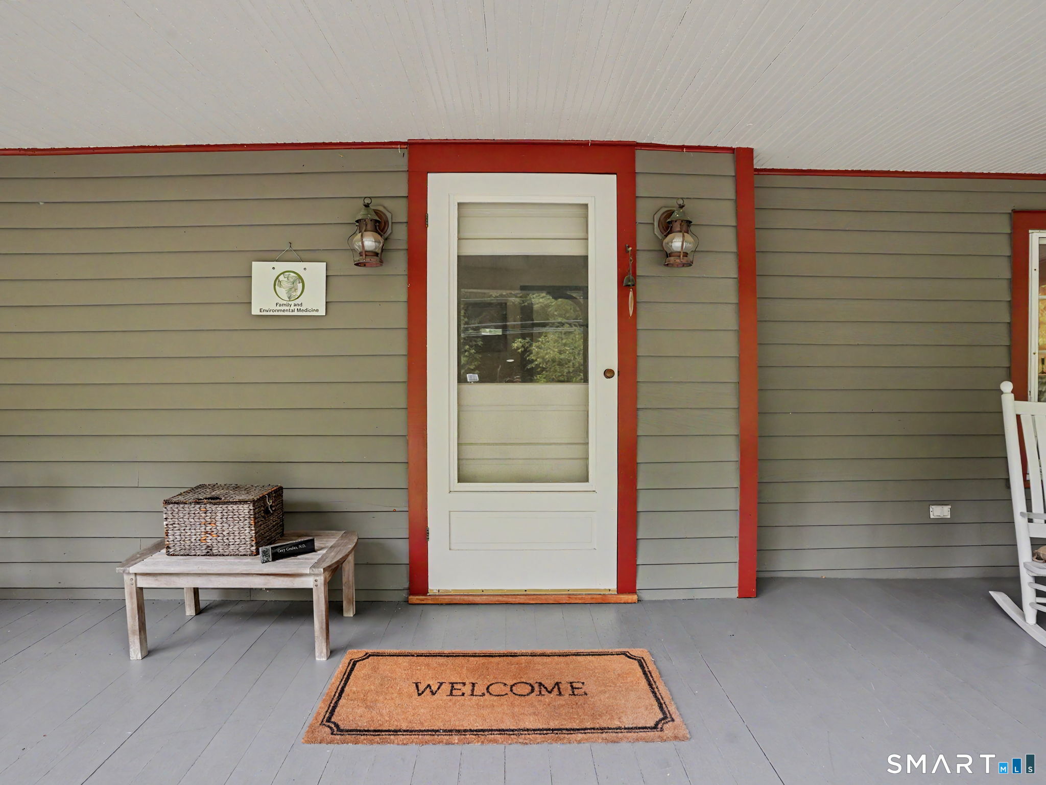 Covered porch entrance with a white door, red trim, gray siding, and a welcome mat.