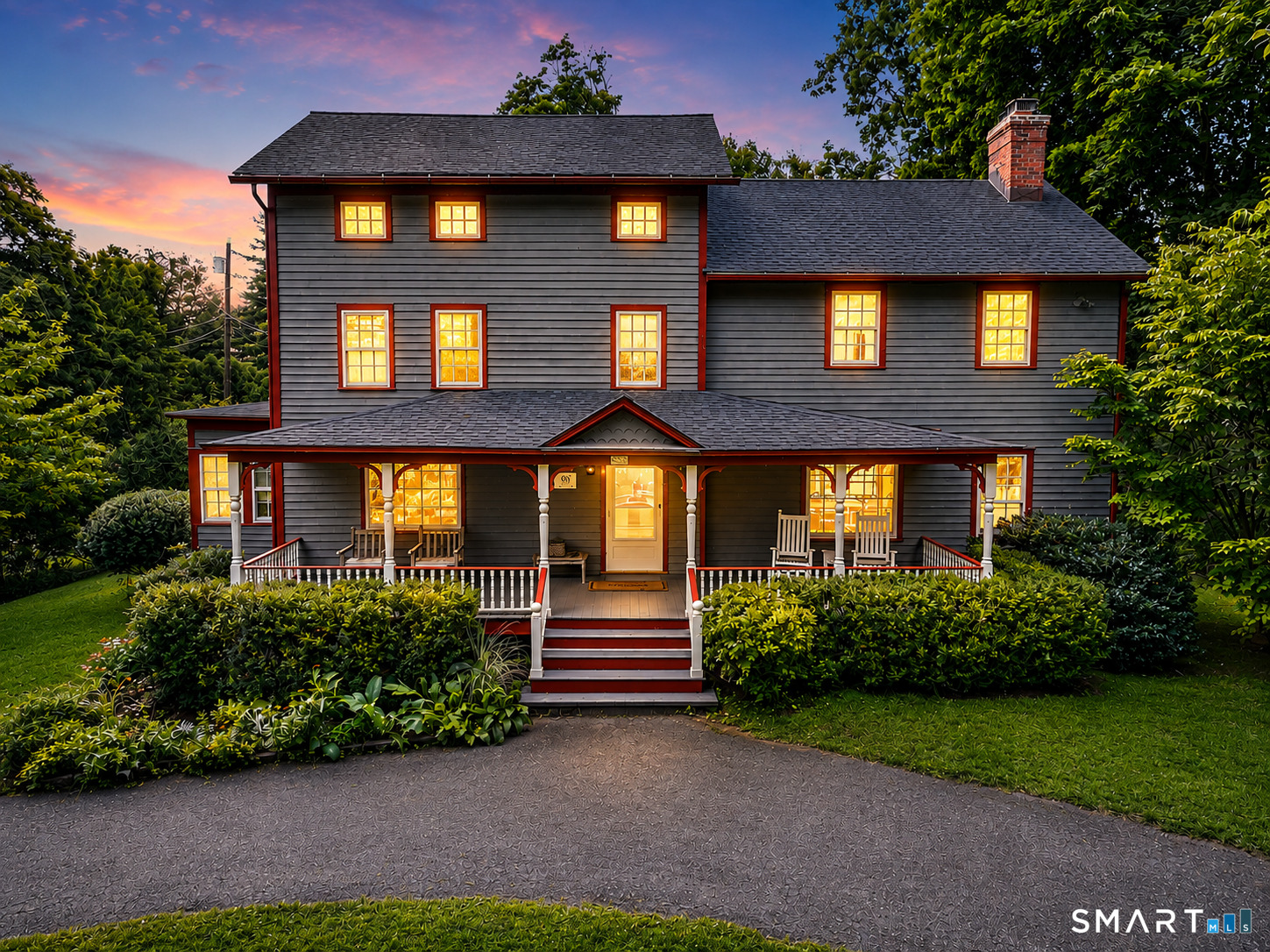 Two-story gray house with red trim, a broad covered porch, and warmly lit windows at dusk.