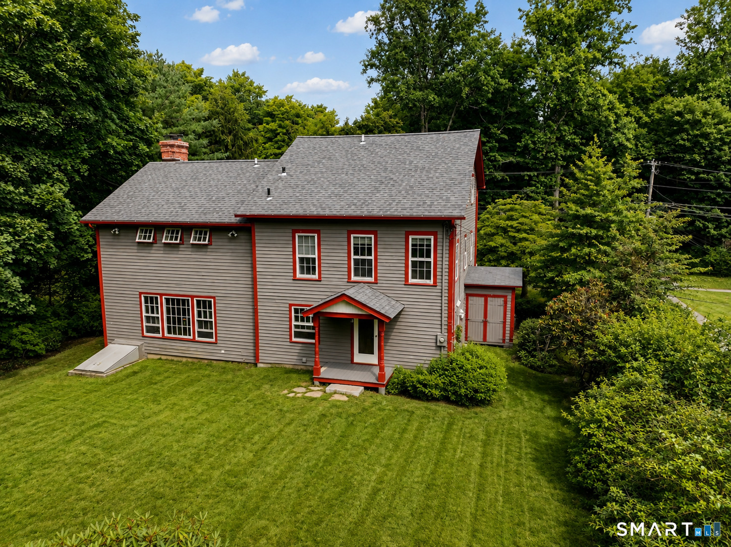 Two-story gray house with red trim, a covered entry, and a broad lawn framed by mature trees.