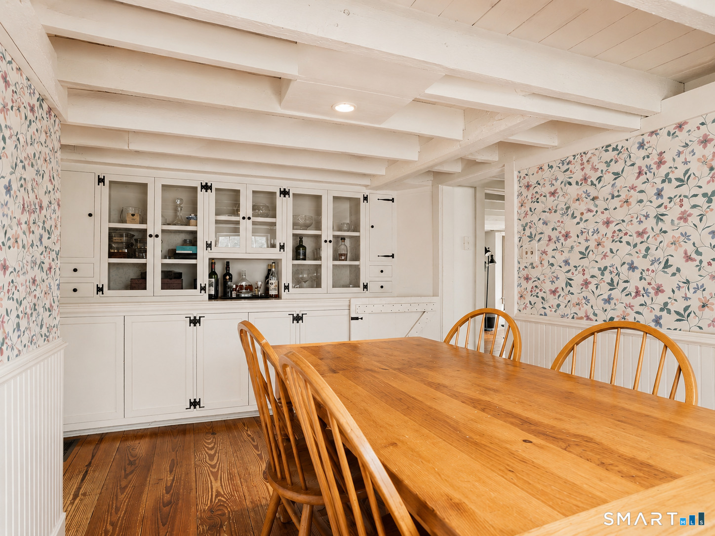 Dining room with a long wooden table, white built-in cabinetry, exposed beams, and floral wallpaper.