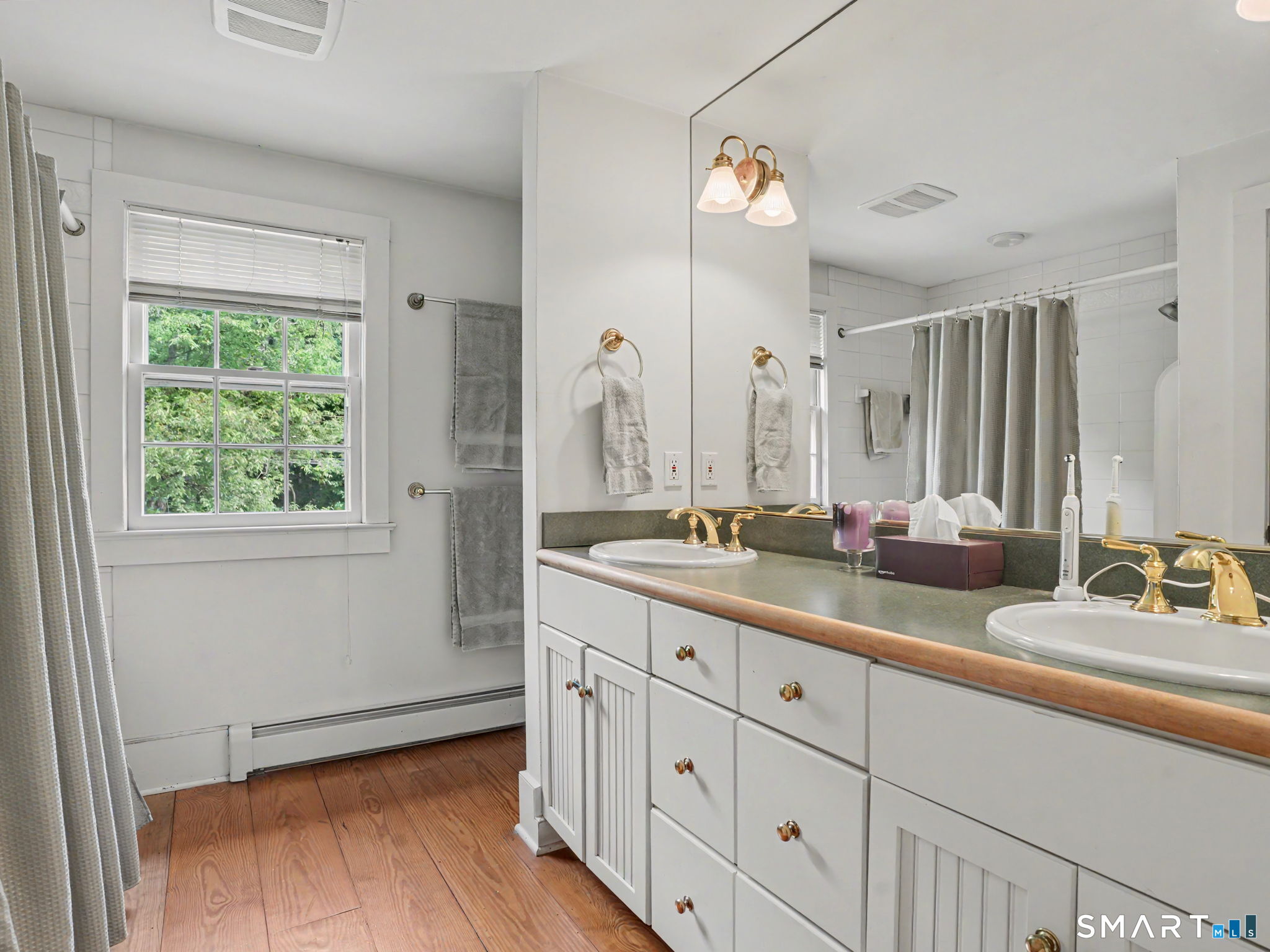 Bathroom with a long double-sink vanity, gold-tone faucets, and a window overlooking greenery.