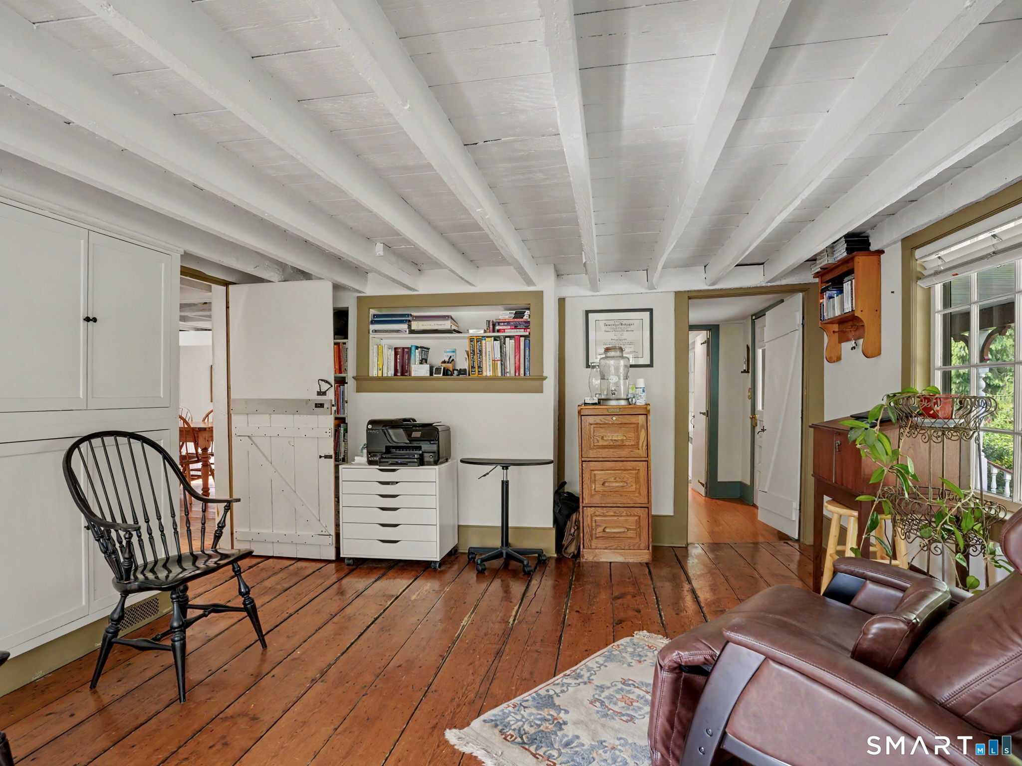 Home office with exposed white beams, wide-plank floors, built-in bookshelves, and a leather sofa.