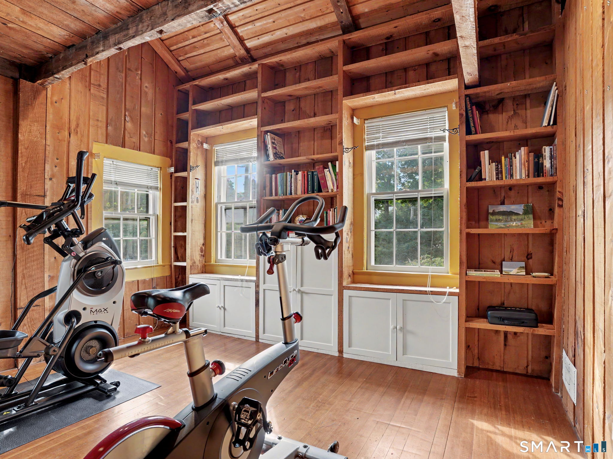 Rustic home gym with stationary bikes, built-in bookshelves, and three sunlit windows.