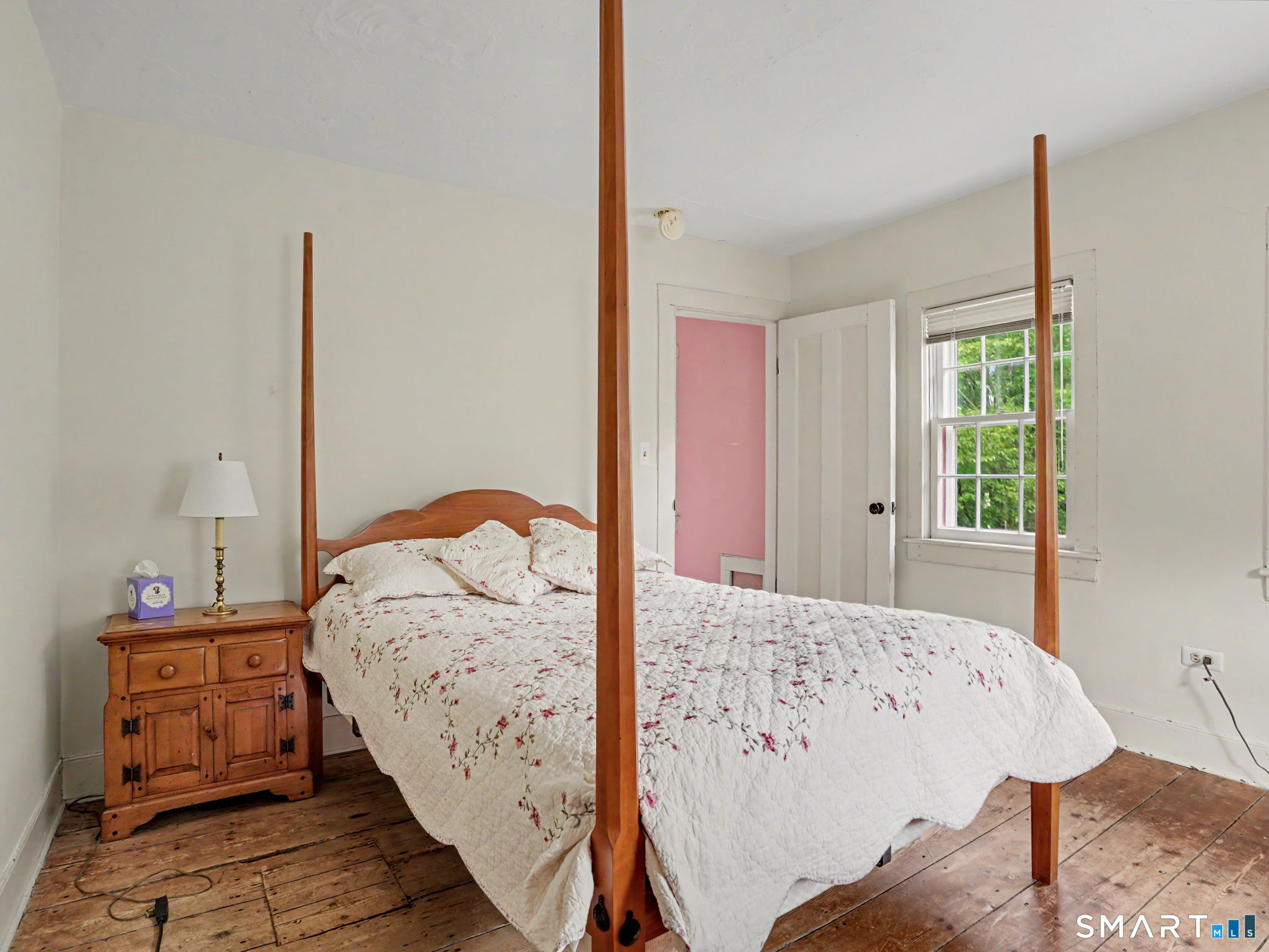 Bedroom with a four-poster bed, floral quilt, rustic nightstand, and pink door beside a leafy window.