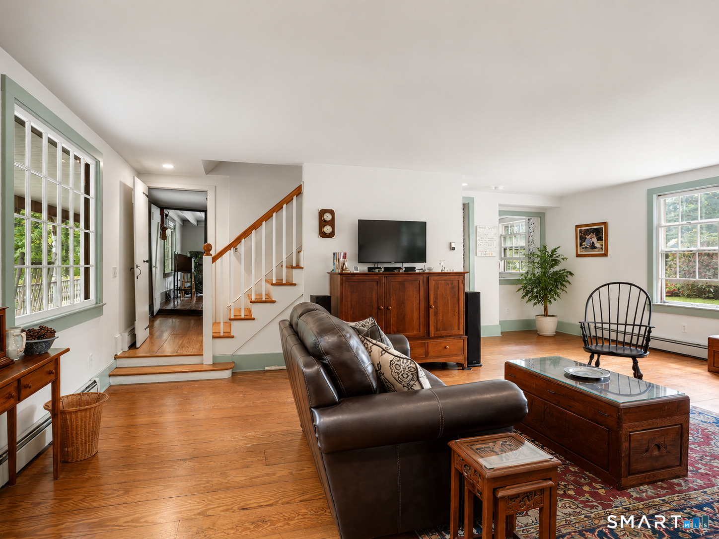 Living room with a leather sofa, polished wood floors, and a staircase beside large windows.