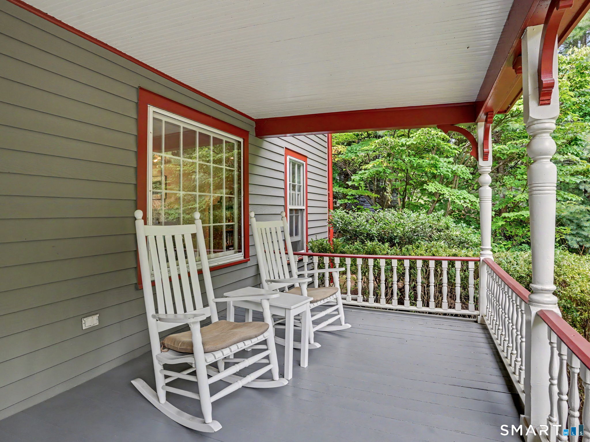 Covered porch with white rocking chairs, gray siding, red trim, and leafy surroundings.