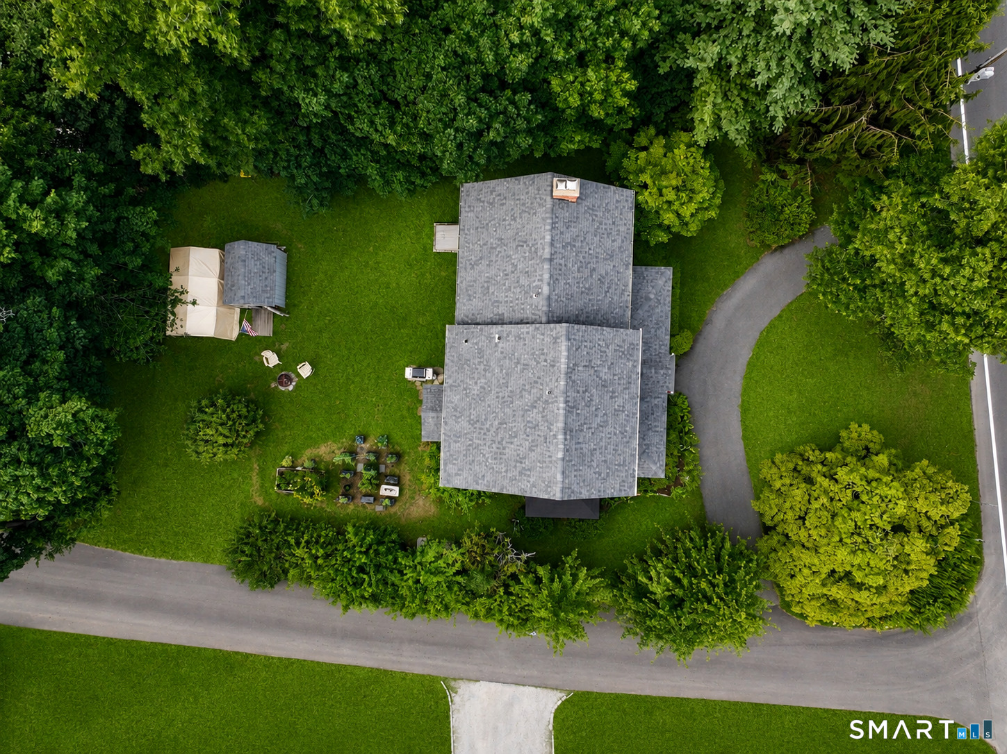 Aerial view of a house surrounded by trees, with a curved driveway, lawn, and garden beds.