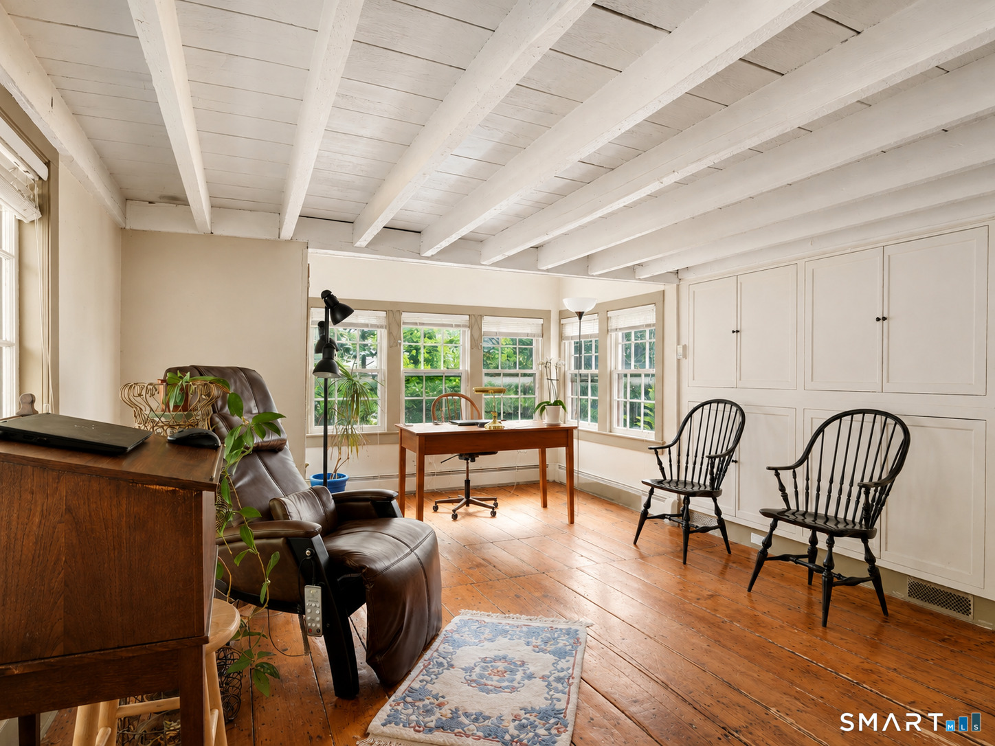 Home office with exposed white beams, built-in cabinetry, and a desk beside bright windows.