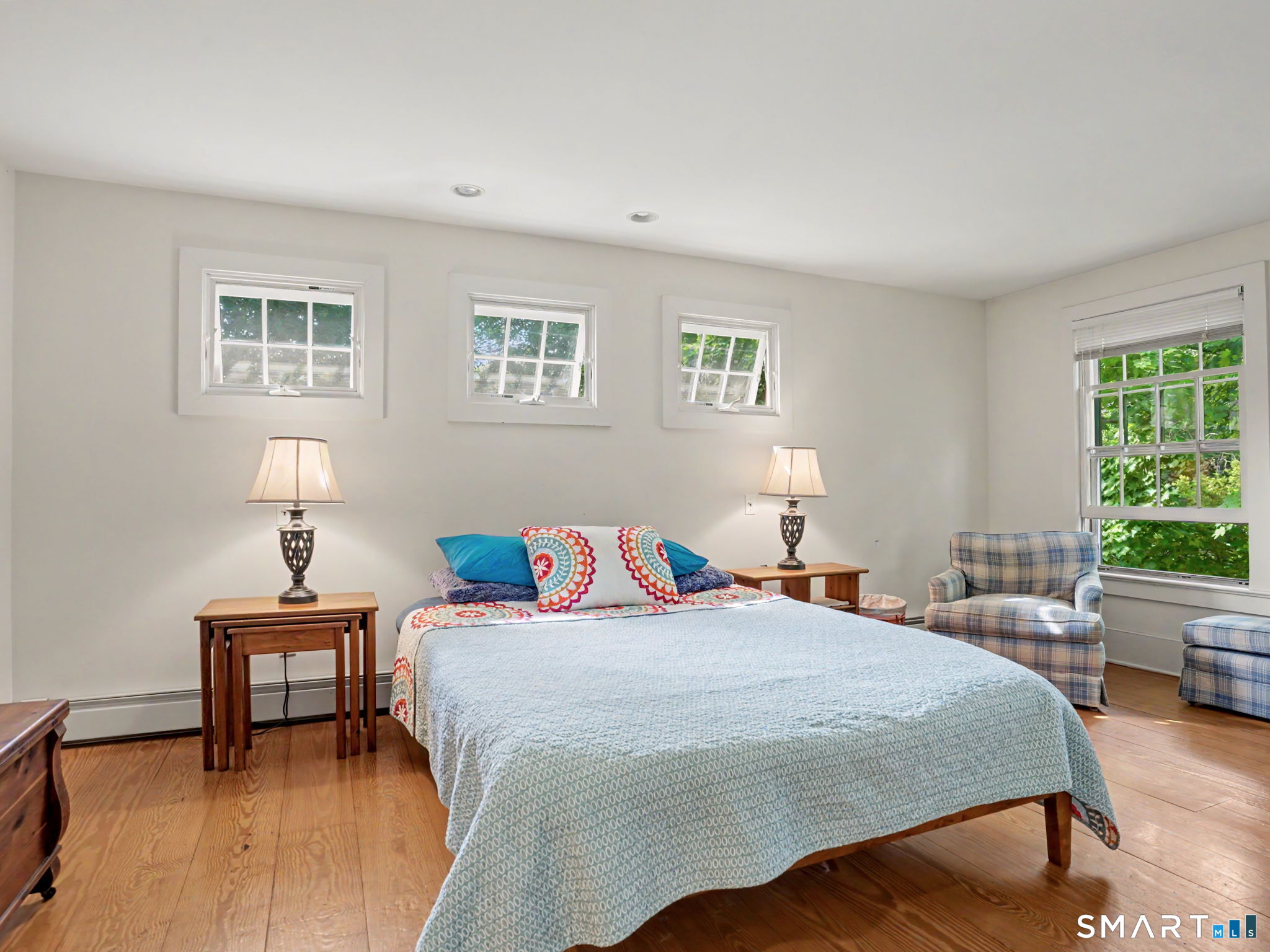 Bedroom with a large pale blue bed, hardwood floors, and windows overlooking greenery.
