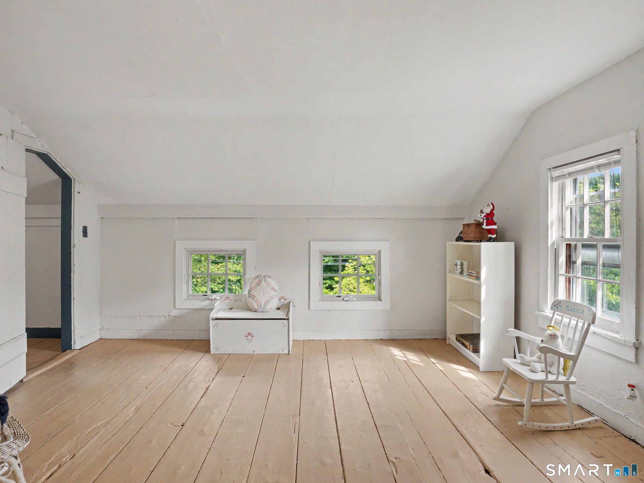 Attic bedroom with sloped ceiling, wide-plank floors, multiple windows, and a child-sized rocking chair.