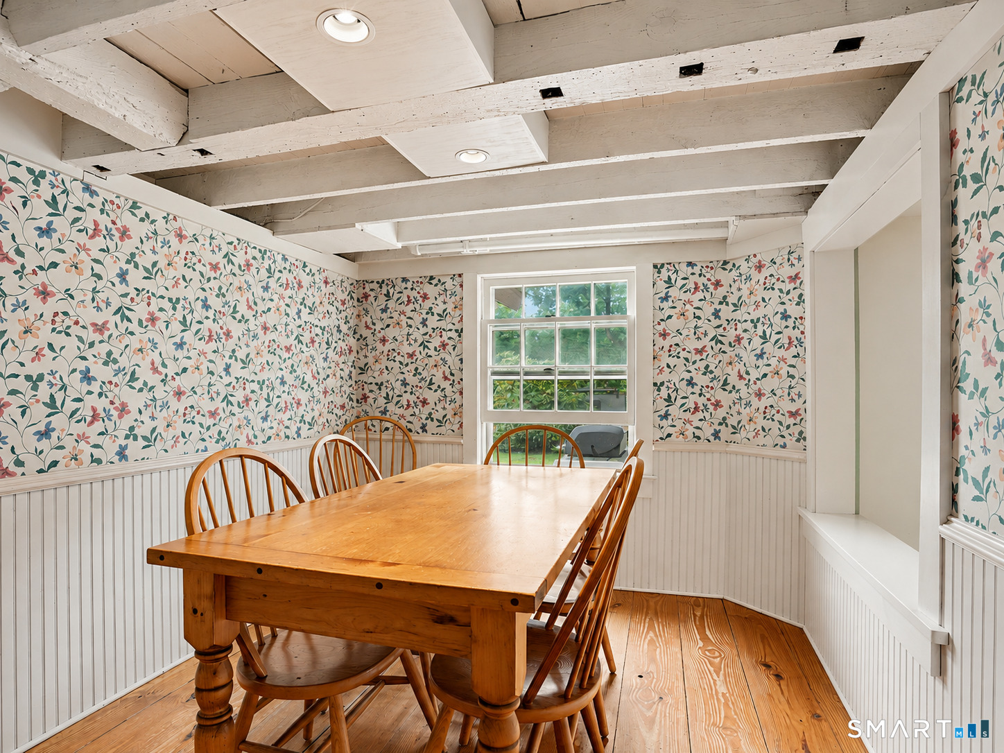 Dining room with a wooden table, floral wallpaper, exposed ceiling beams, and a window overlooking greenery.