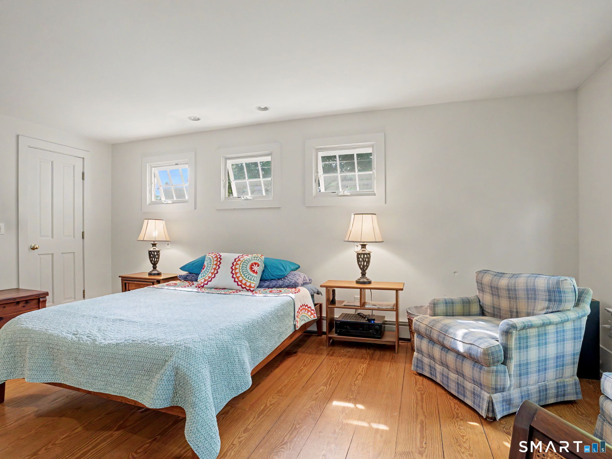 Furnished bedroom with a blue-covered bed, plaid armchair, wood floors, and three small windows.