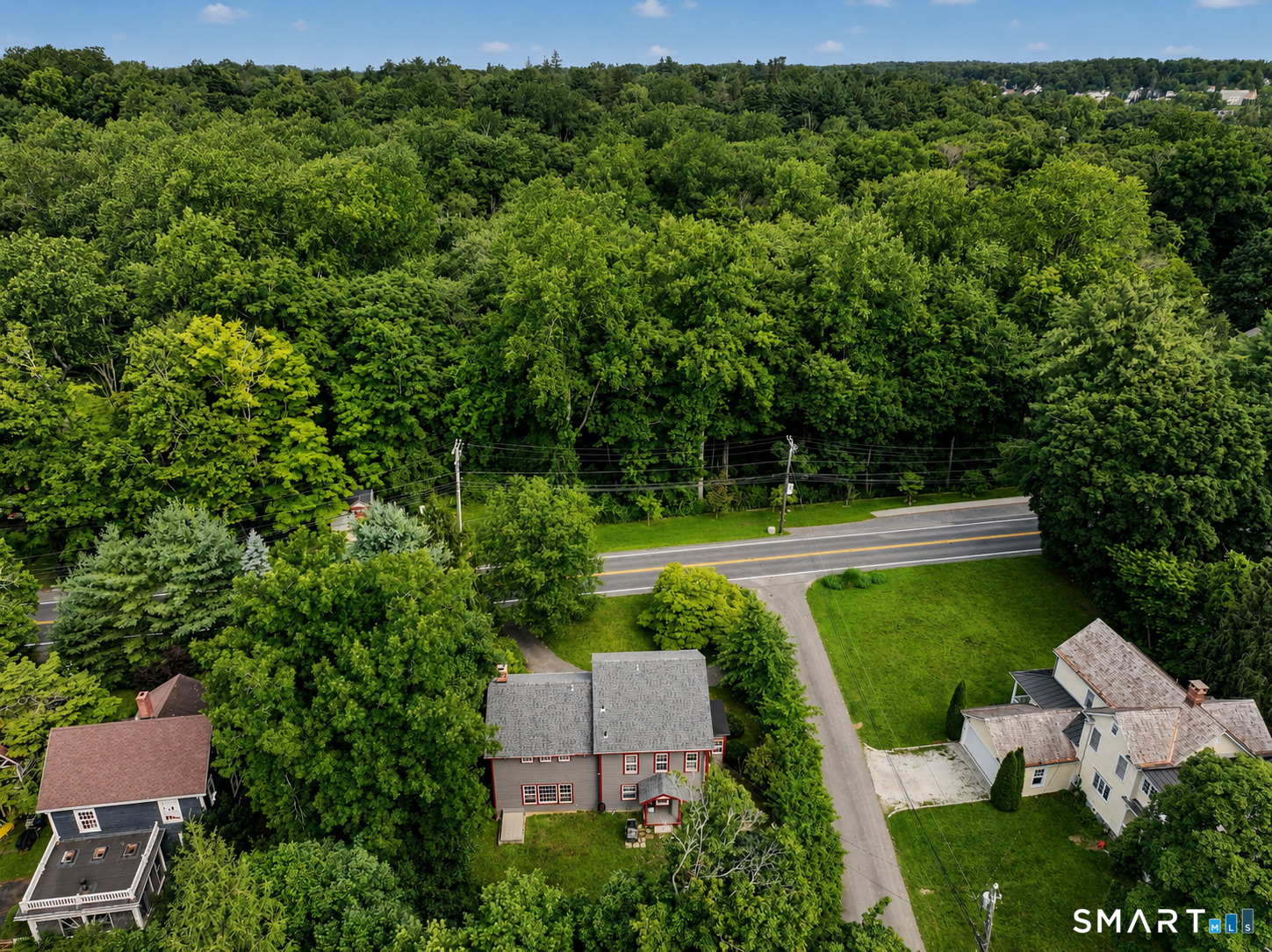 Aerial view of a gray house with red trim beside a road, bordered by lawns and dense woodland.