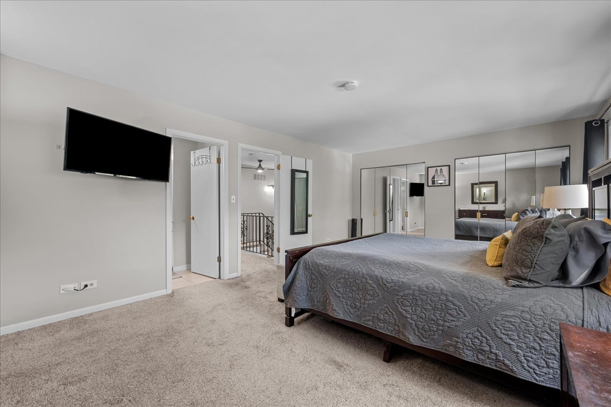 Bedroom with a large gray-quilted bed, mirrored closet doors, and wall-mounted television.