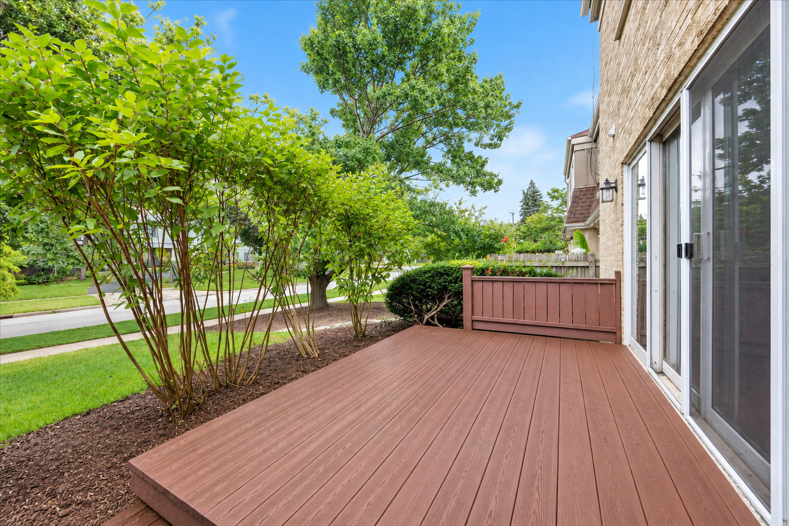 Wooden deck beside a brick home, bordered by leafy shrubs and sliding glass doors.