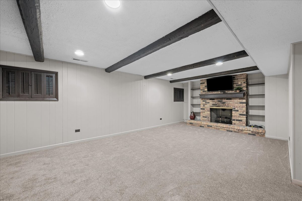 Carpeted lower-level family room with exposed beams and a brick fireplace with built-in shelving.