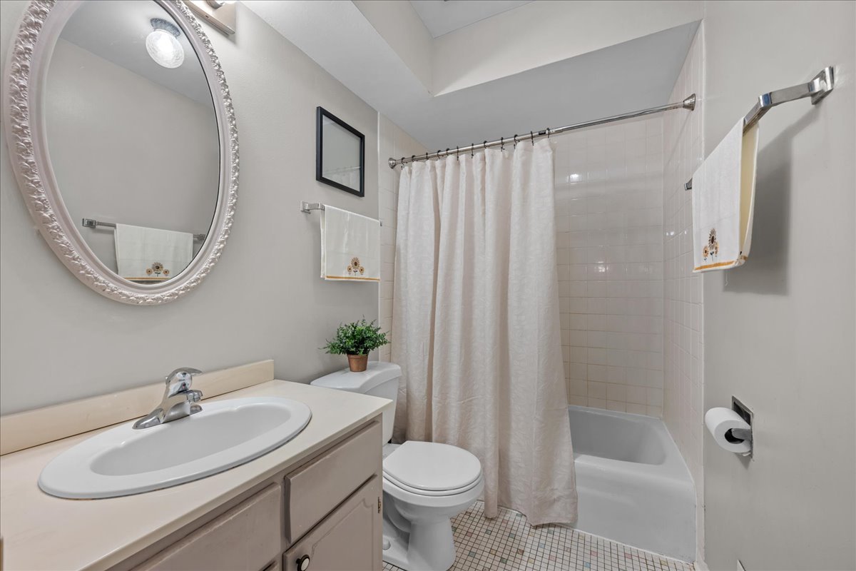 Bathroom with an oval mirror, sink vanity, toilet, and white-curtained bathtub.