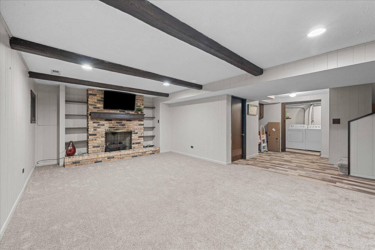 Empty living room with exposed ceiling beams, brick fireplace, built-in shelves, and laundry area beyond.