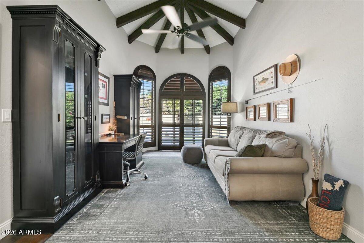 Living room and office with a gray sofa, dark built-ins, arched shuttered windows, and exposed ceiling beams.