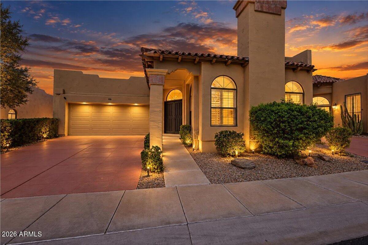 Stucco home at dusk with a two-car garage, arched windows, and illuminated desert landscaping.