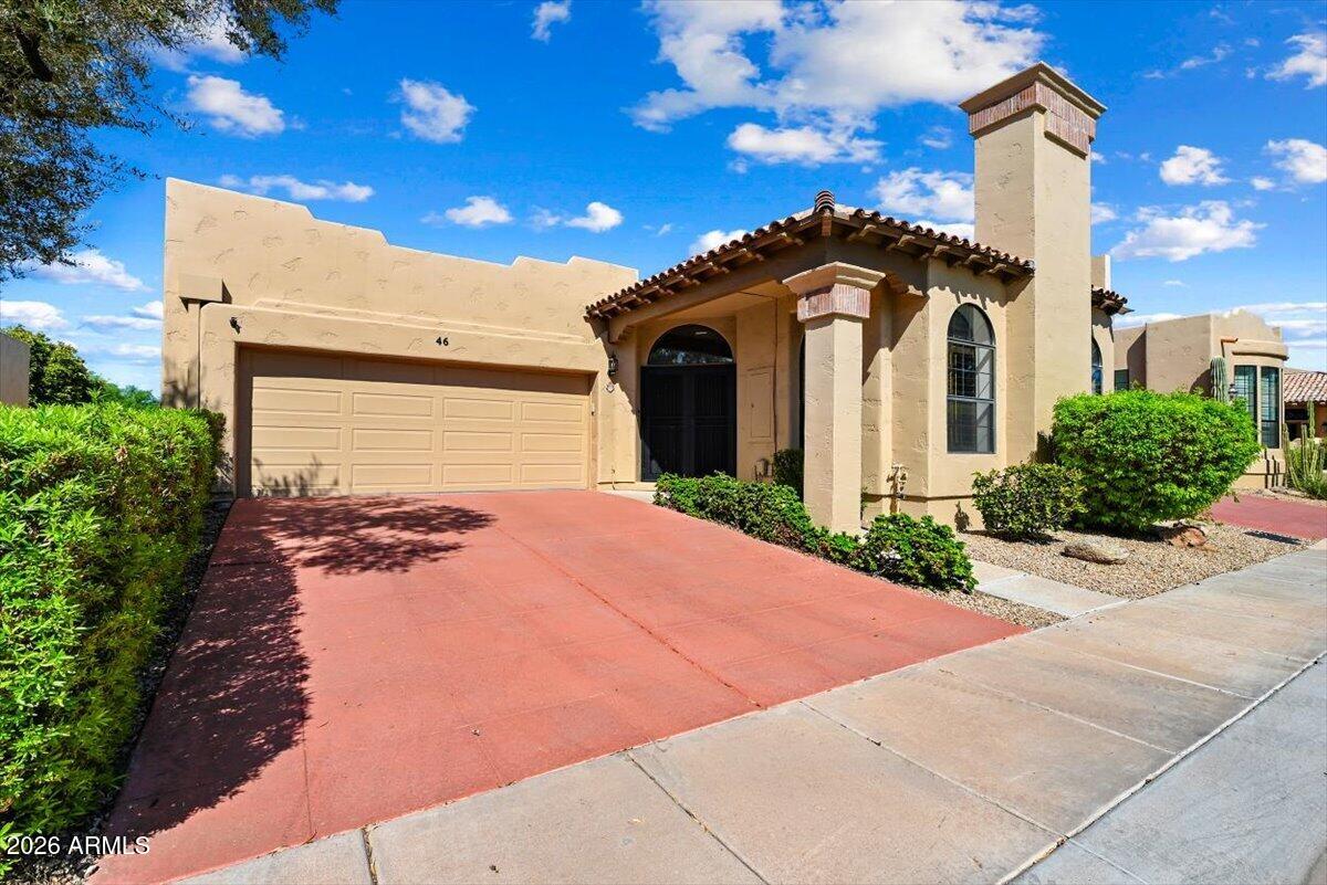 Tan stucco home with a red driveway, two-car garage, arched entry, and tall chimney.