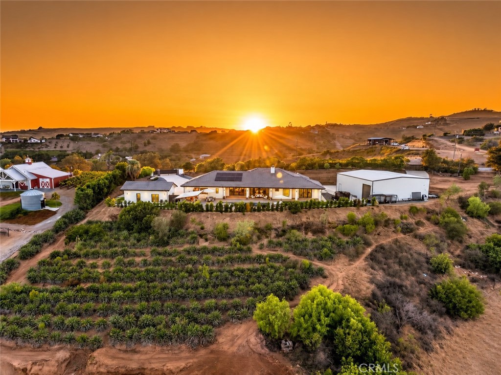 Aerial view of a rural home and planted rows at sunset, with solar panels and outbuildings.
