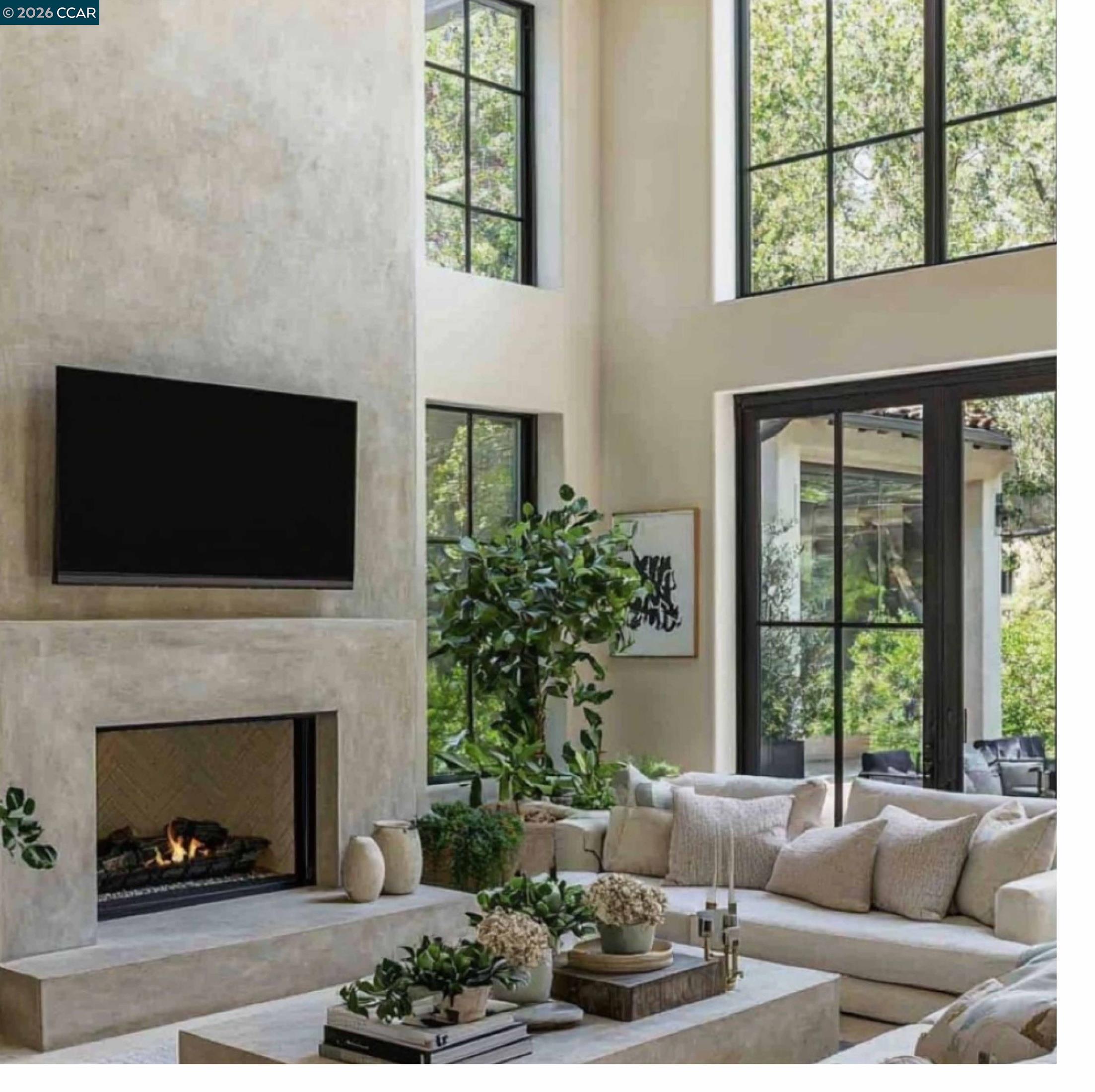 Double-height living room with a stone fireplace, cream sectional, and tall windows overlooking trees.