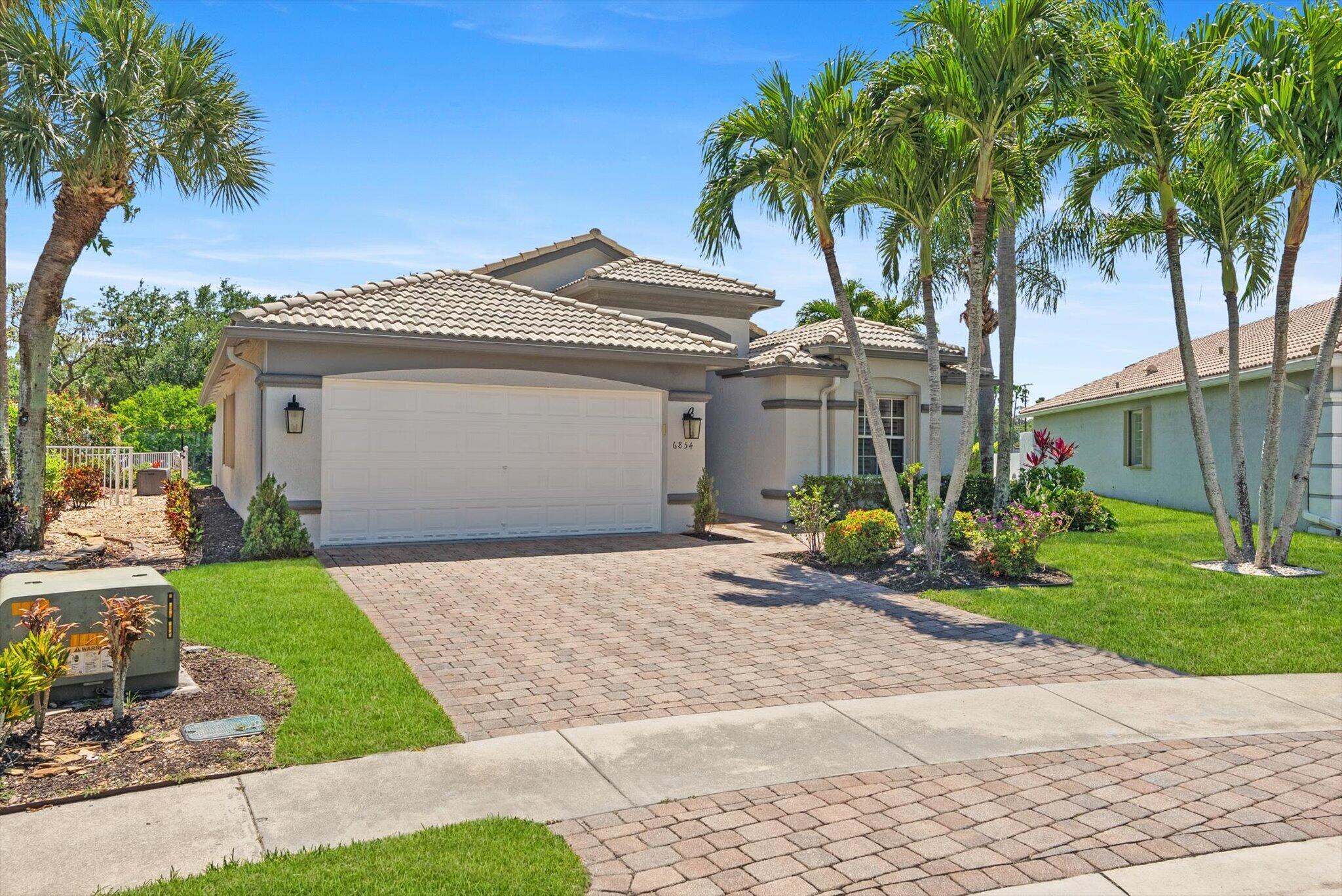 Single-story home with a tiled roof, two-car garage, brick-paver driveway, and palm trees.