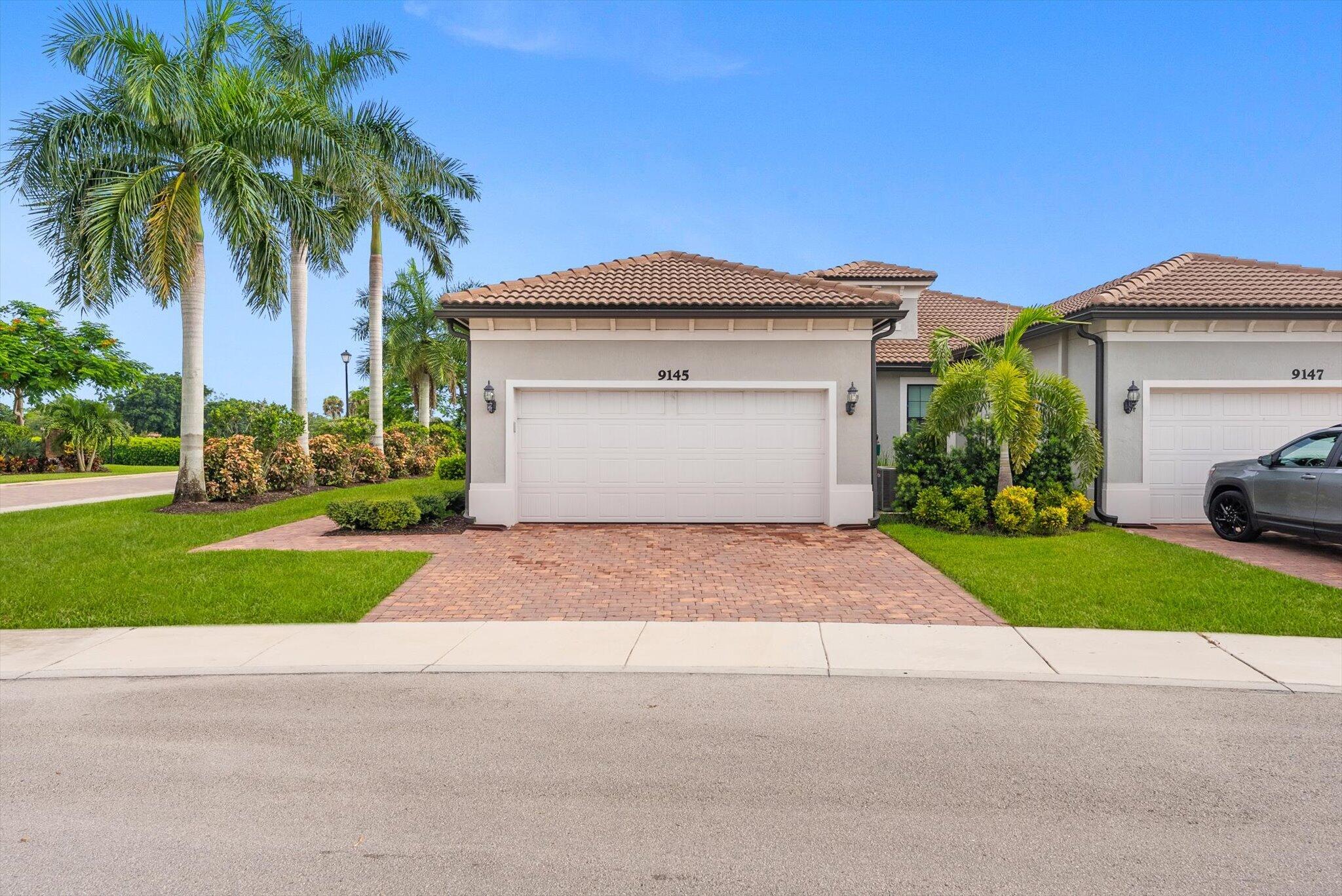 Home exterior with a white double garage, brick-paver driveway, and tall palm trees.